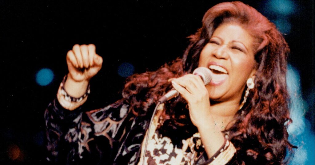 Live performance photo of Aretha Franklin singing passionately into a microphone, wearing a patterned long coat.