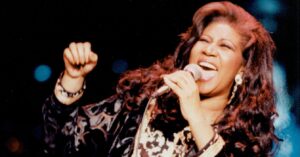 Live performance photo of Aretha Franklin singing passionately into a microphone, wearing a patterned long coat.