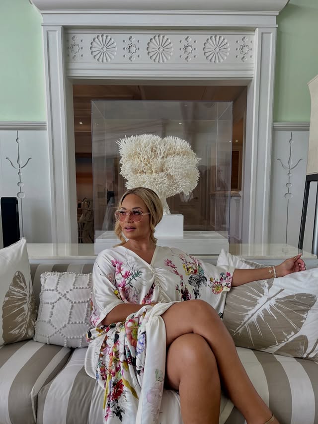 Gizelle Bryant seated on a striped sofa in a floral robe, wearing sunglasses with decorative coral display behind her.