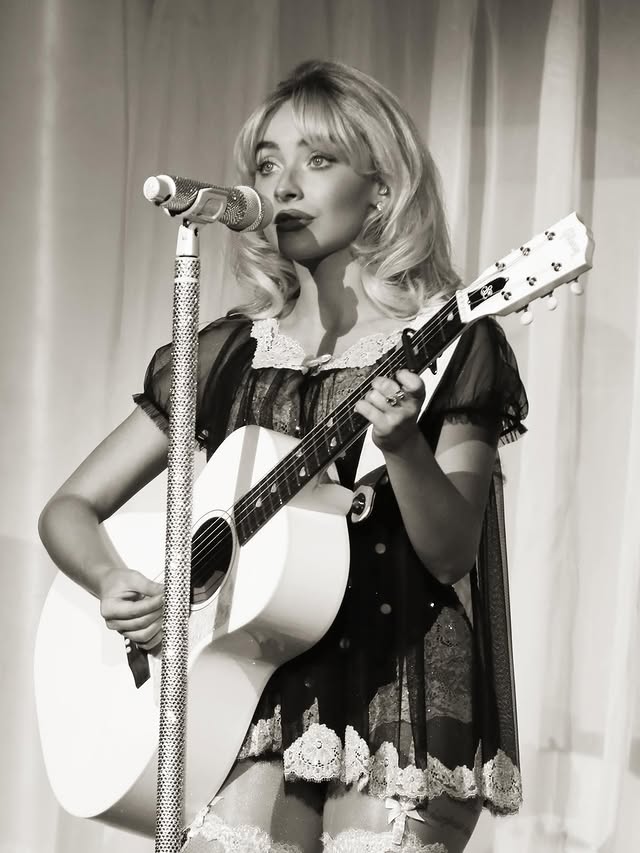 Write Alt Text (for accessibility) Alt Text: Singer Sabrina Carpenter on stage in a country-western outfit, wearing a gingham top, sequined shorts, and cowboy boots, holding a microphone.