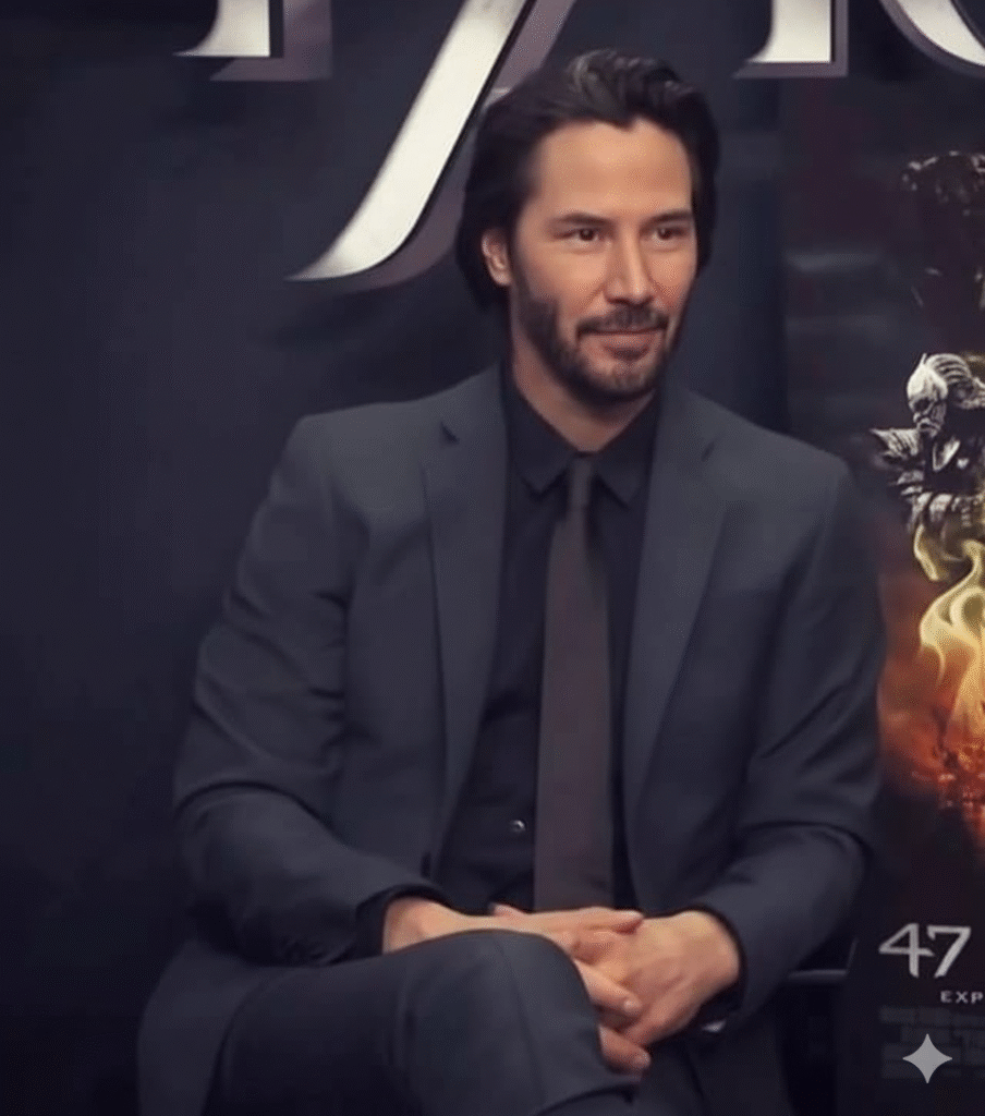 A portrait of actor Keanu Reeves seated, wearing a dark gray suit, black shirt, and tie, smiling subtly against a dark promotional backdrop.