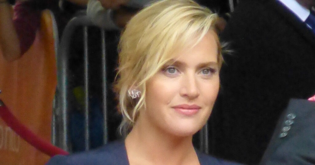 Young Kate Winslet in a 90s-style photoshoot, wearing a black leather jacket and red lipstick, looking intently at the camera.