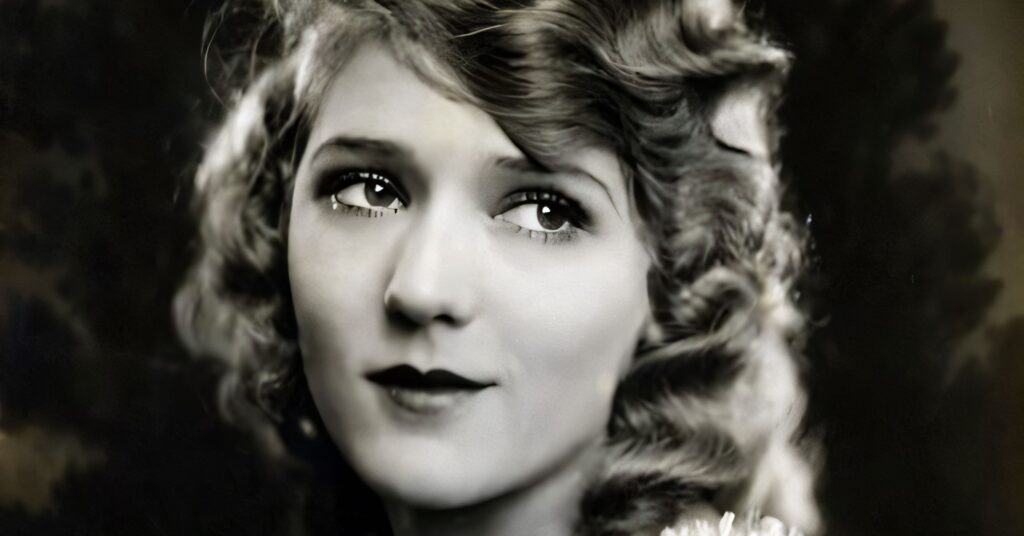 A sepia-toned close-up portrait of Mary Pickford with her iconic long ringlet curls, looking upward with a soft expression and wearing a ruffled lace-trimmed garment.