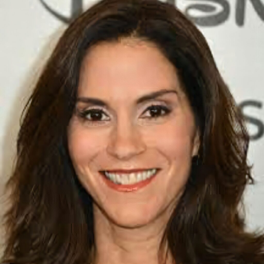 A woman with short, dark, textured hair smiles, wearing a black strapless top and a sequined shrug with a heart pendant necklace.