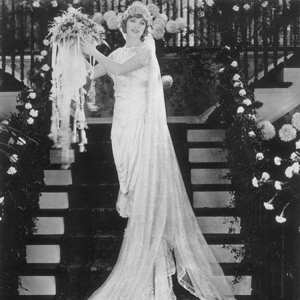 A vintage black-and-white full-length photograph of a bride in a 1920s wedding gown, standing on a floral-decorated staircase and holding a large bouquet with long ribbons.