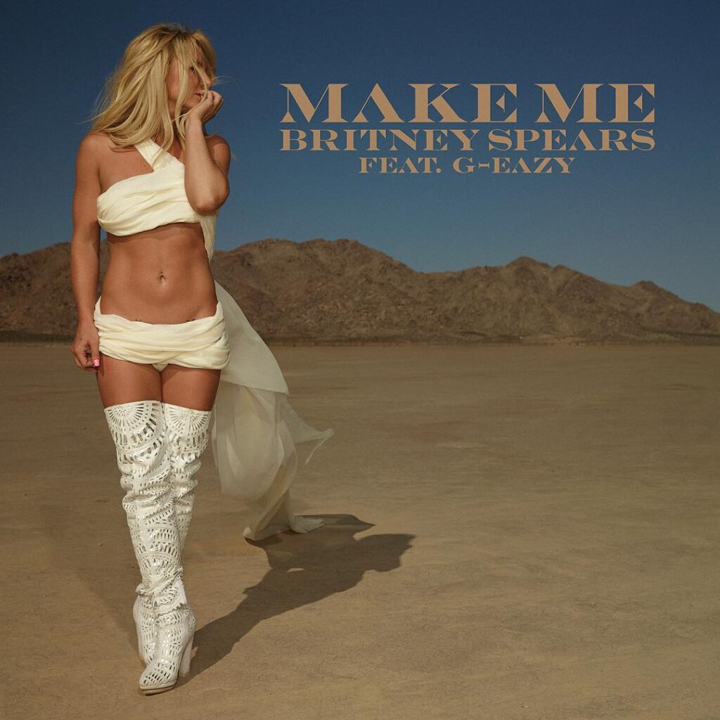 Britney Spears' "Make Me..." single cover. She is standing in a desert, wearing a white, flowing cloth outfit and knee-high lace-up white boots.