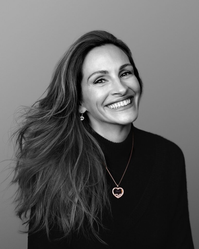 A candid, side-profile photo of actress Julia Roberts smiling widely. She has wavy blonde hair and is wearing a dark blazer over a black T-shirt with the word "DAVE" repeated three times in stacked red, orange, and yellow retro lettering.