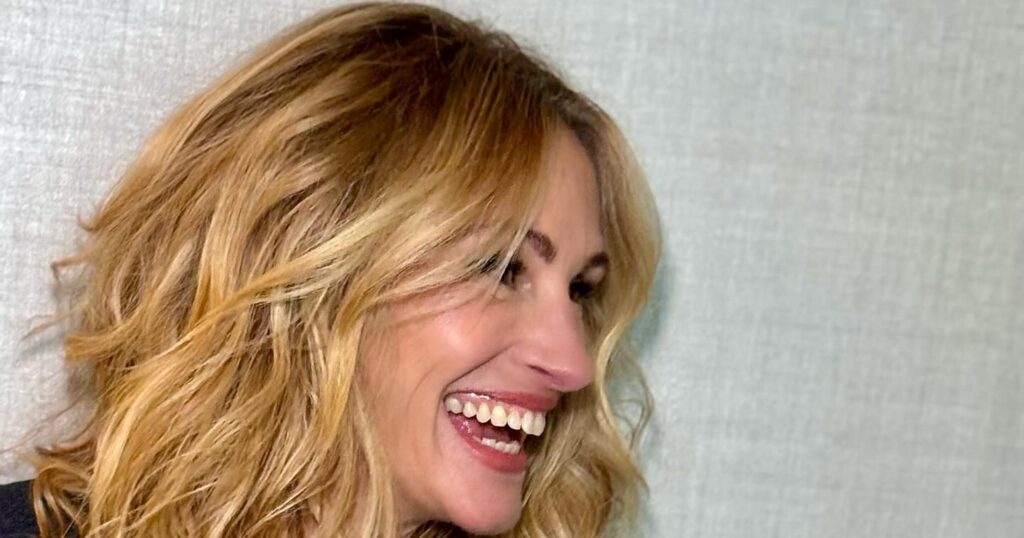A black and white portrait of actress Julia Roberts smiling directly at the camera. Her long, dark hair flows to the side, and she wears a dark top with a diamond-studded heart pendant necklace.