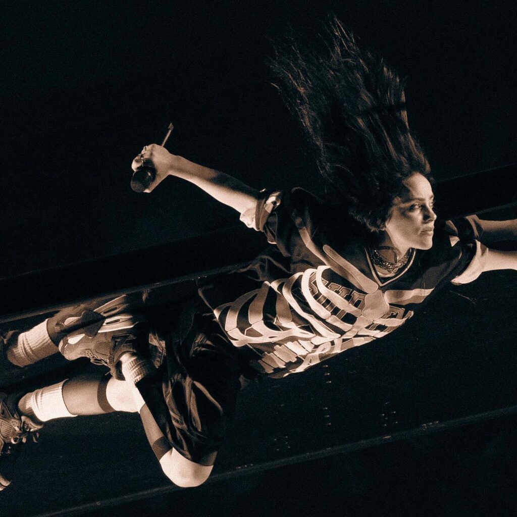 Black and white photo of Billie Eilish mid-air, leaning back with her long dark hair flowing. She wears a skeleton-print t-shirt and shorts while holding a microphone.