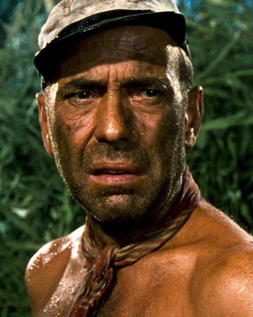 A color close-up of Humphrey Bogart as Charlie Allnut in The African Queen, appearing sweaty, dirty, and distressed against a jungle background.