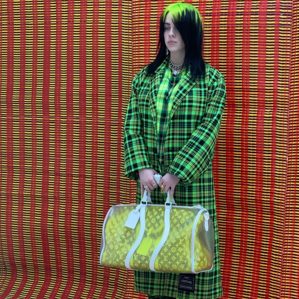 Billie Eilish stands against a red and yellow woven mat background. She wears a bright neon green plaid coat and holds a large yellow Louis Vuitton duffel bag.