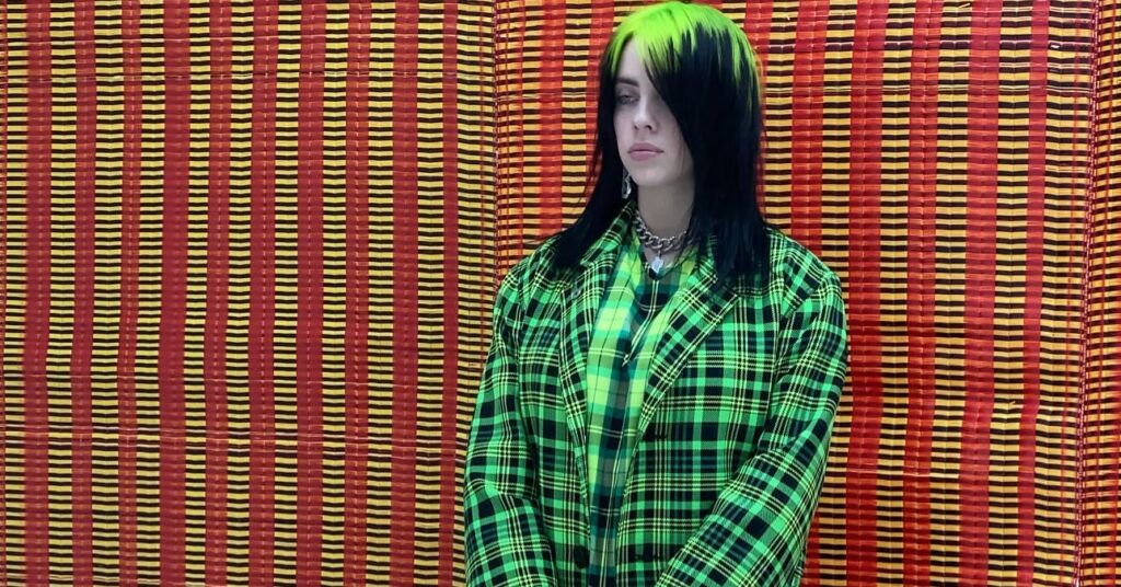 Billie Eilish stands against a red and yellow woven mat background. She wears a bright neon green plaid coat and holds a large yellow Louis Vuitton duffel bag.