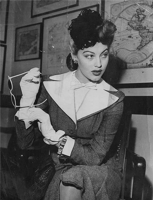 A vintage black-and-white photograph of actress Ava Gardner sitting in a chair, wearing a tailored suit with a white bow tie, white gloves, and holding sunglasses.