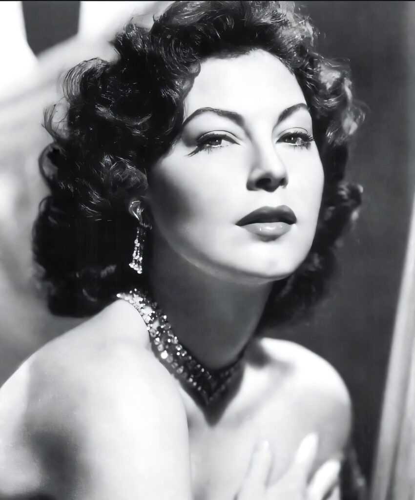 A close-up black-and-white studio portrait of Ava Gardner featuring her signature dark wavy hair, arched eyebrows, and a sparkling diamond necklace and earrings.