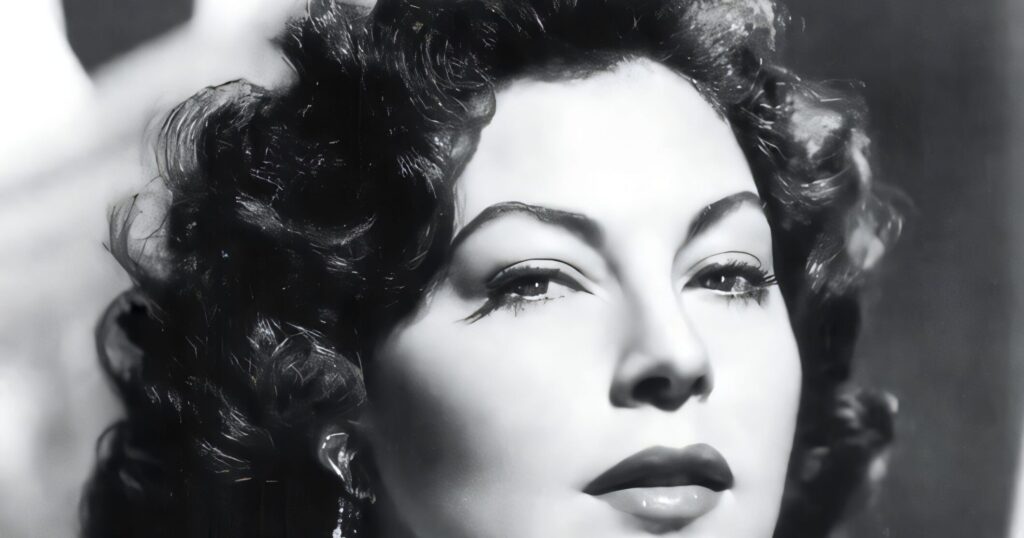 A close-up black-and-white studio portrait of Ava Gardner featuring her signature dark wavy hair, arched eyebrows, and a sparkling diamond necklace and earrings.