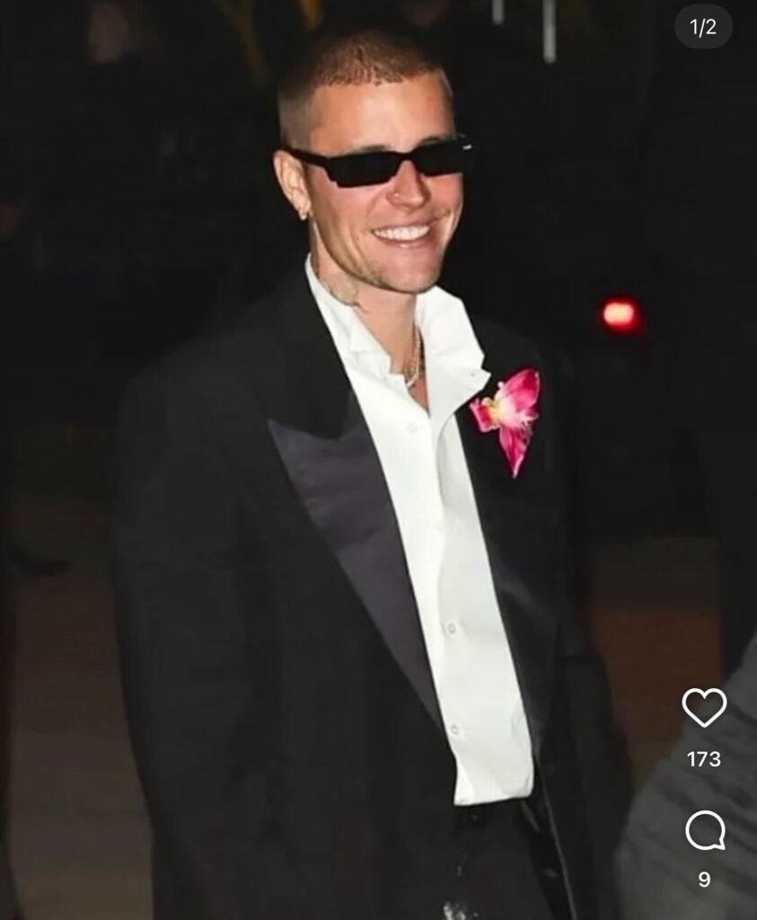 Justin Bieber wearing a black tuxedo jacket with satin lapels, a white collared shirt, black sunglasses, and a large, bright pink floral lapel pin, smiling brightly outdoors at night.