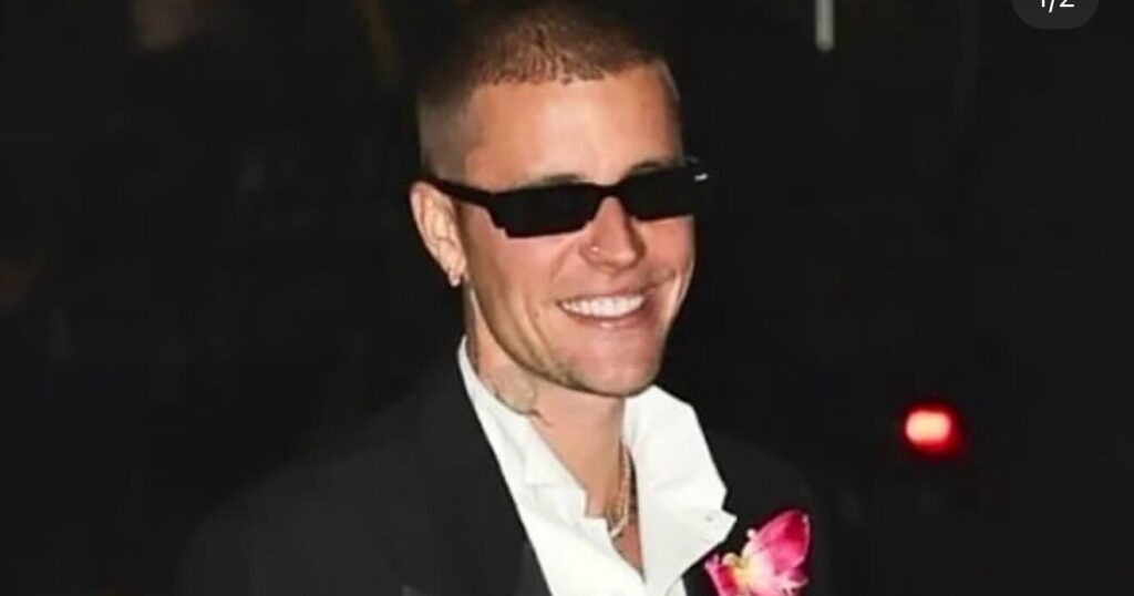 Justin Bieber wearing a black tuxedo jacket with satin lapels, a white collared shirt, black sunglasses, and a large, bright pink floral lapel pin, smiling brightly outdoors at night.