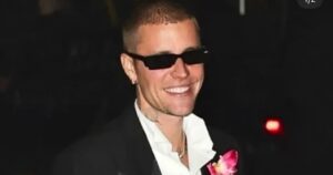 Justin Bieber wearing a black tuxedo jacket with satin lapels, a white collared shirt, black sunglasses, and a large, bright pink floral lapel pin, smiling brightly outdoors at night.