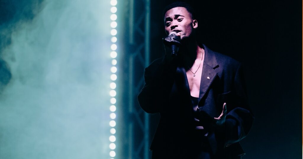A male singe Denzel Washingtonr in a long dark coat performs onstage under dramatic blue lighting and fog, holding a microphone with one hand while gesturing with the other.