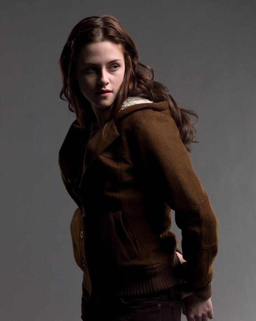 Actress Kristen Stewart with long, wavy brown hair, wearing a cozy brown wool jacket with a shearling-lined collar, looking over her shoulder against a neutral gray background.