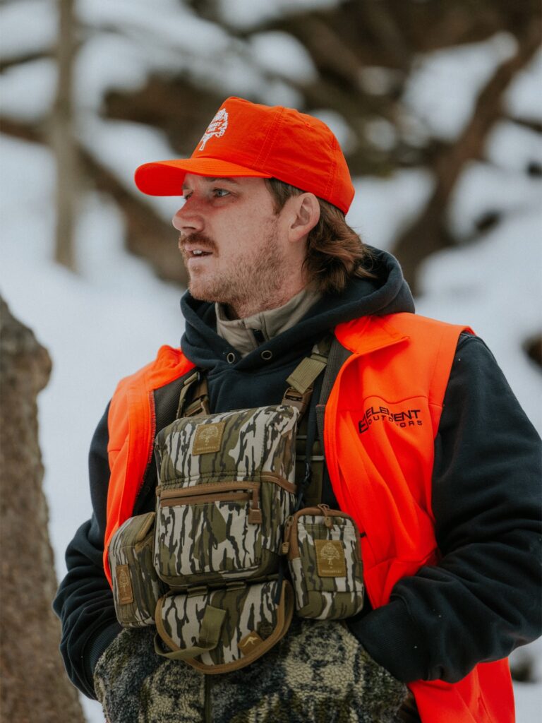 Morgan Wallen outdoors in a snowy setting, wearing bright neon orange hunting gear, a black hoodie, and a camouflage chest rig pack from Marsupial Gear, topped with a vibrant orange cap.