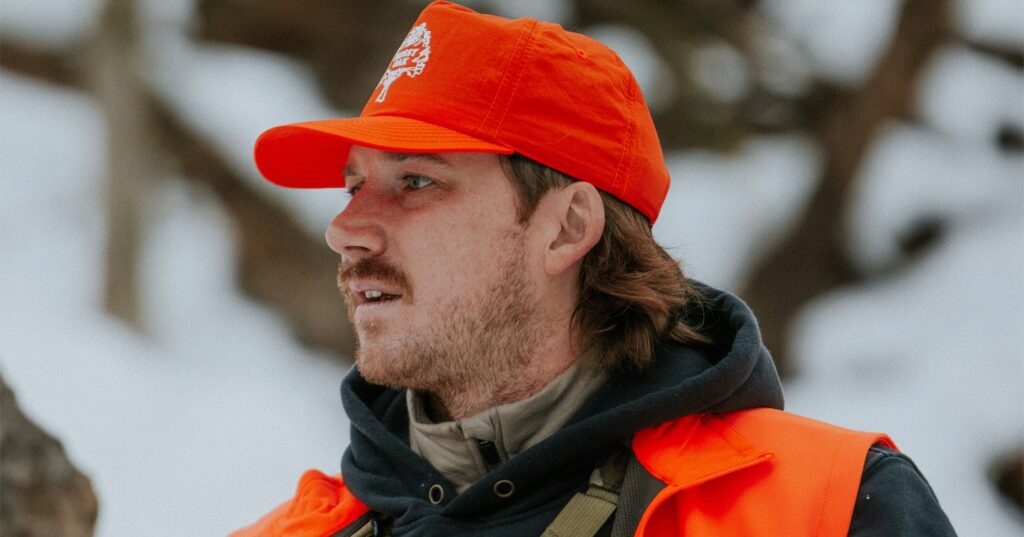 Morgan Wallen outdoors in a snowy setting, wearing bright neon orange hunting gear, a black hoodie, and a camouflage chest rig pack from Marsupial Gear, topped with a vibrant orange cap.