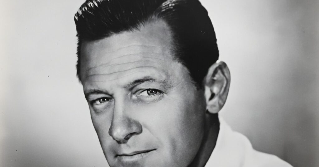 A colorized, upscaled vintage photograph of a young, smiling man in a brown tweed blazer, white shirt, and patterned red tie. He has reddish-brown hair and blue eyes, set against a soft blue and lavender studio backdrop.