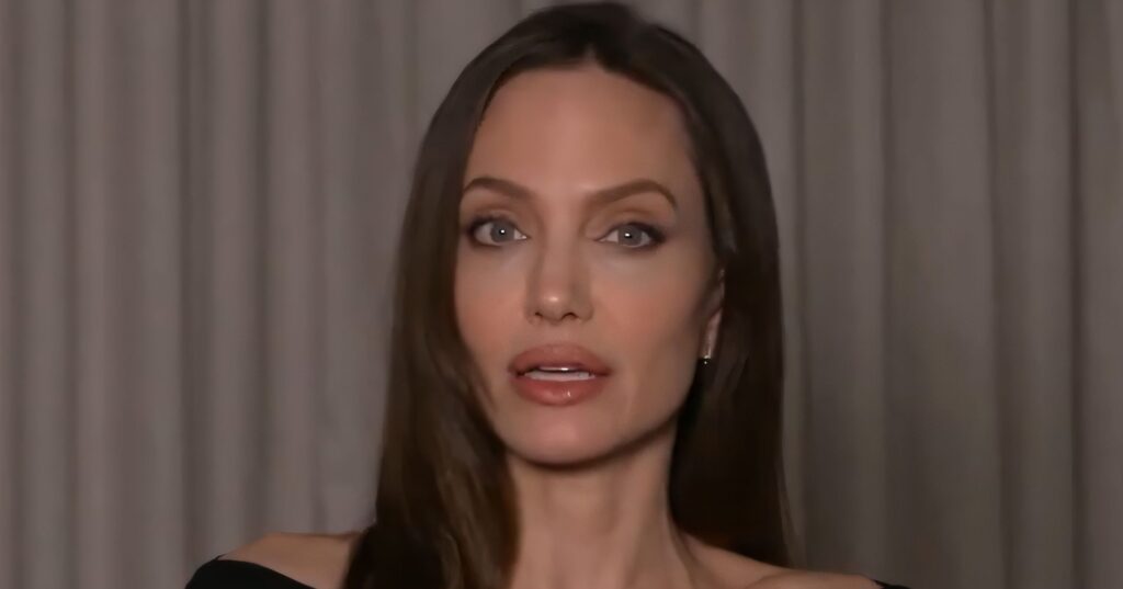 Close-up of Angelina Jolie, a middle-aged woman with long brown hair, wearing a black off-the-shoulder top against a neutral, curtained background. She is looking directly at the camera with a neutral expression.