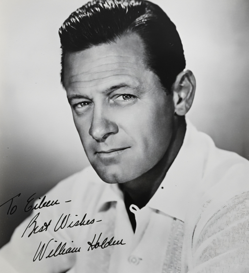 A colorized, upscaled vintage photograph of a young, smiling man in a brown tweed blazer, white shirt, and patterned red tie. He has reddish-brown hair and blue eyes, set against a soft blue and lavender studio backdrop.