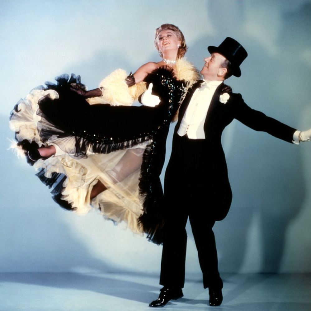 Fred Astaire in a tuxedo and top hat lifting Ginger Rogers, who wears an elegant ruffled gown.