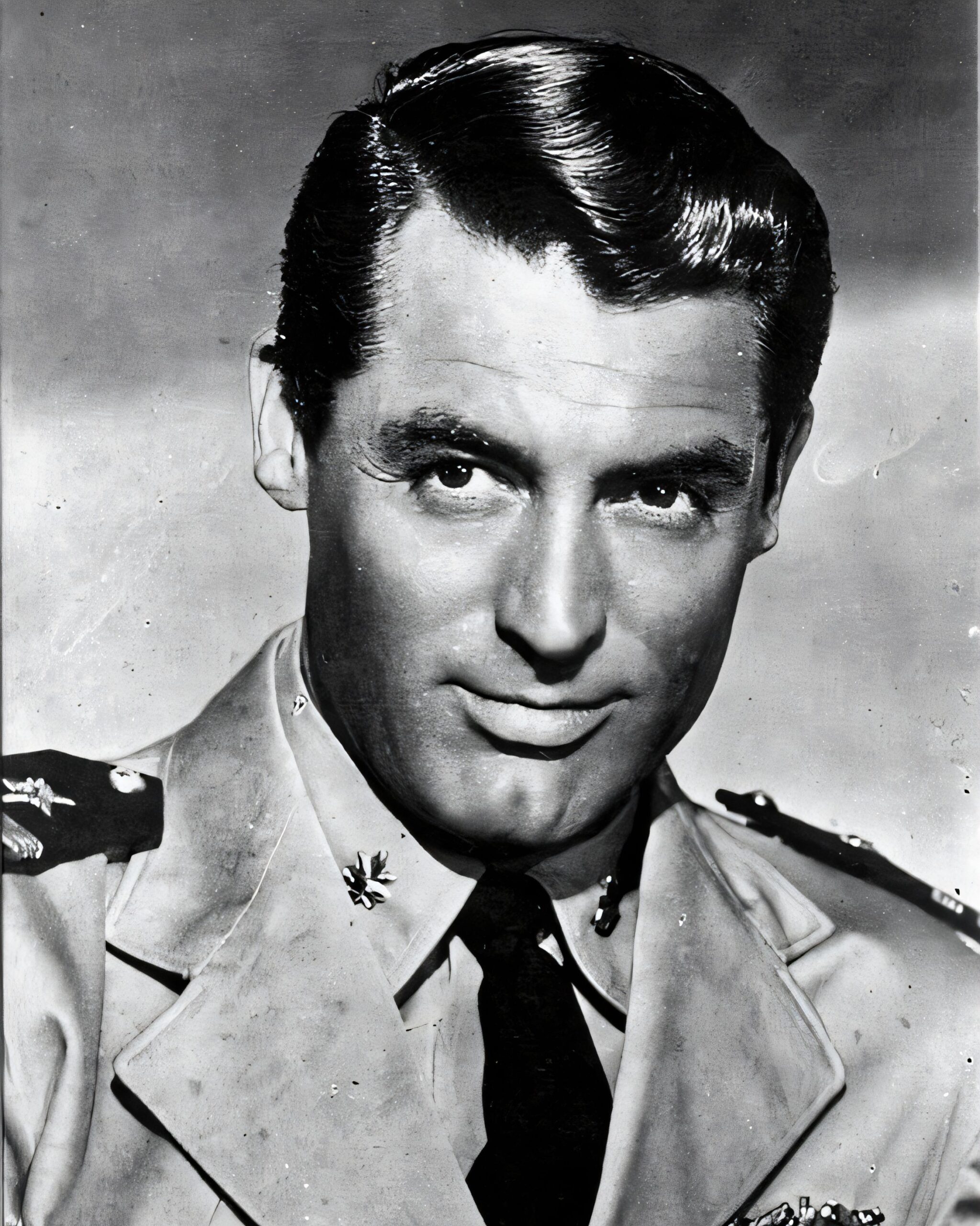 A close-up color photograph of Cary Grant’s face, showing his tan complexion, dark hair with slight graying at the temples, and intense brown eyes.