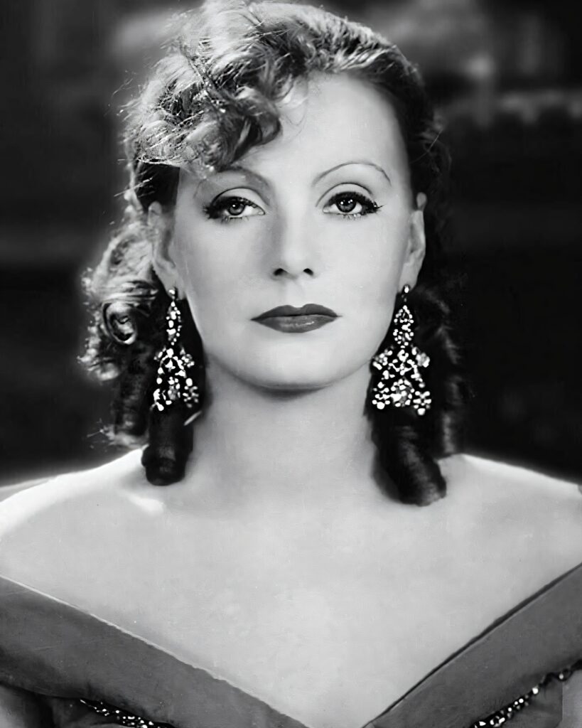 A high-contrast black and white close-up portrait of Greta Garbo looking directly at the camera with a neutral expression.