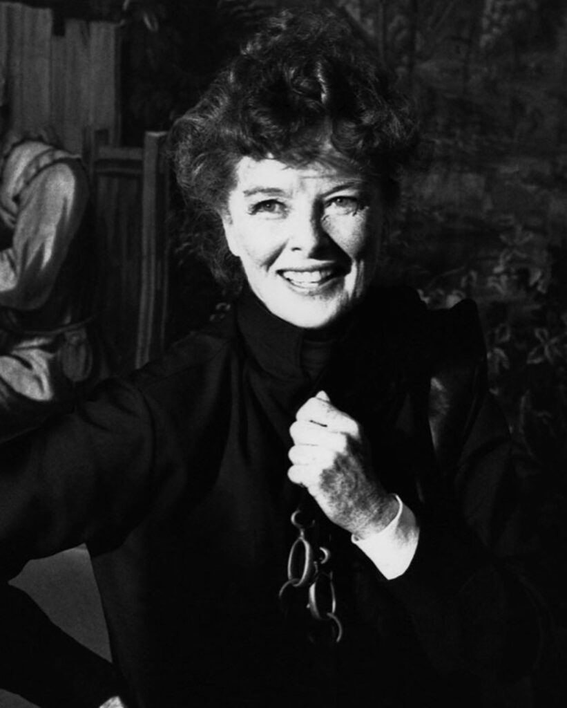 A monochromatic portrait of actress Katharine Hepburn smiling warmly, featuring her signature curly hair and freckled complexion, while wearing a dark, high-collared garment.