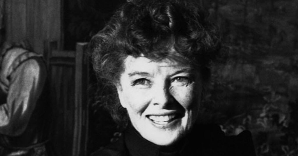 A monochromatic portrait of actress Katharine Hepburn smiling warmly, featuring her signature curly hair and freckled complexion, while wearing a dark, high-collared garment.