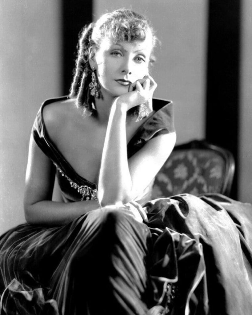 A black and white full-length photograph of Greta Garbo seated in an ornate gown, resting her chin thoughtfully on her hand.