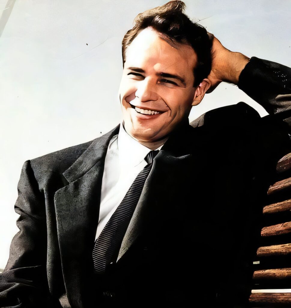 A color photograph of a young, smiling Marlon Brando wearing a grey suit, white shirt, and dark patterned tie.