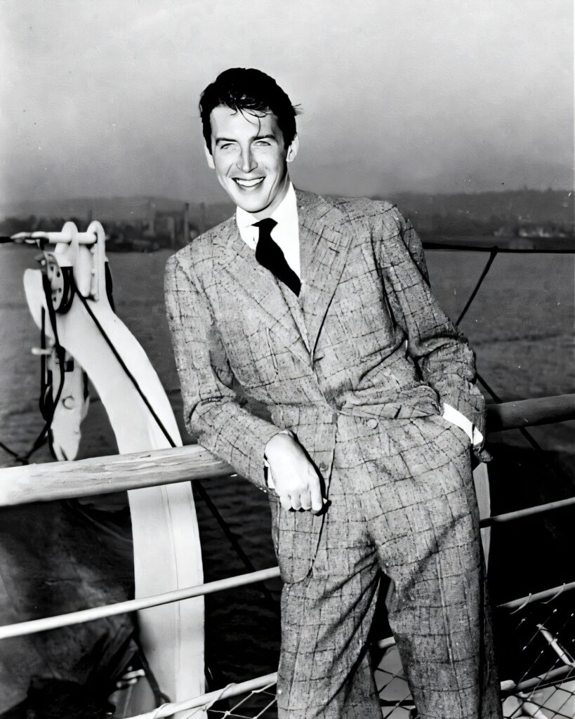 A vintage black and white photograph of a young, smiling man in a patterned suit leaning against a ship's railing.