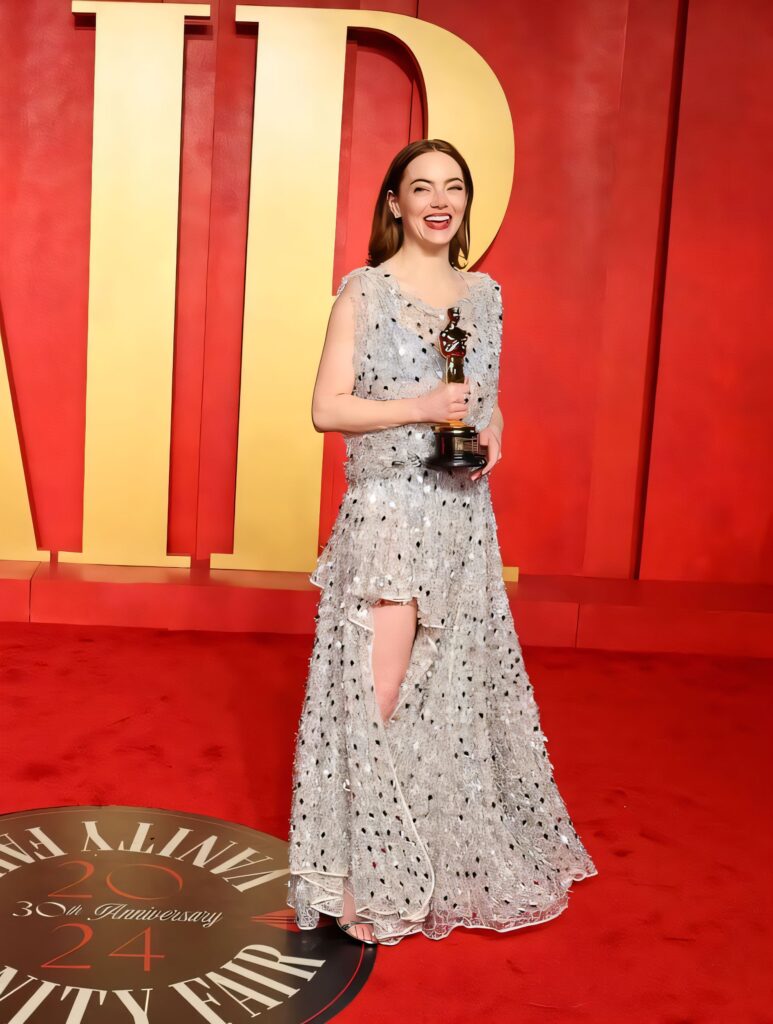 Write the Alt Text Emma Stone smiling broadly on a red carpet, holding an Oscar statuette while wearing a sparkling, sheer silver floor-length gown.