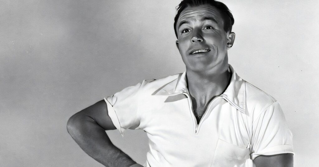 A high-contrast black and white studio portrait of Gene Kelly posing with one leg raised and hands on his hips.