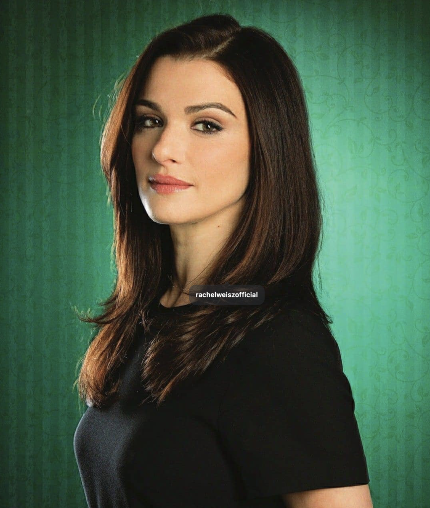 A medium close-up shot of a man with a dark, thick beard and mustache looking directly at the camera with a serious expression. He has dark, swept-back hair and is wearing a black t-shirt with a large blue and white graphic on the front. He is positioned in the foreground against a vibrant, out-of-focus city street at night, filled with bright neon signs, glowing building lights, and the blurred streaks of passing cars.