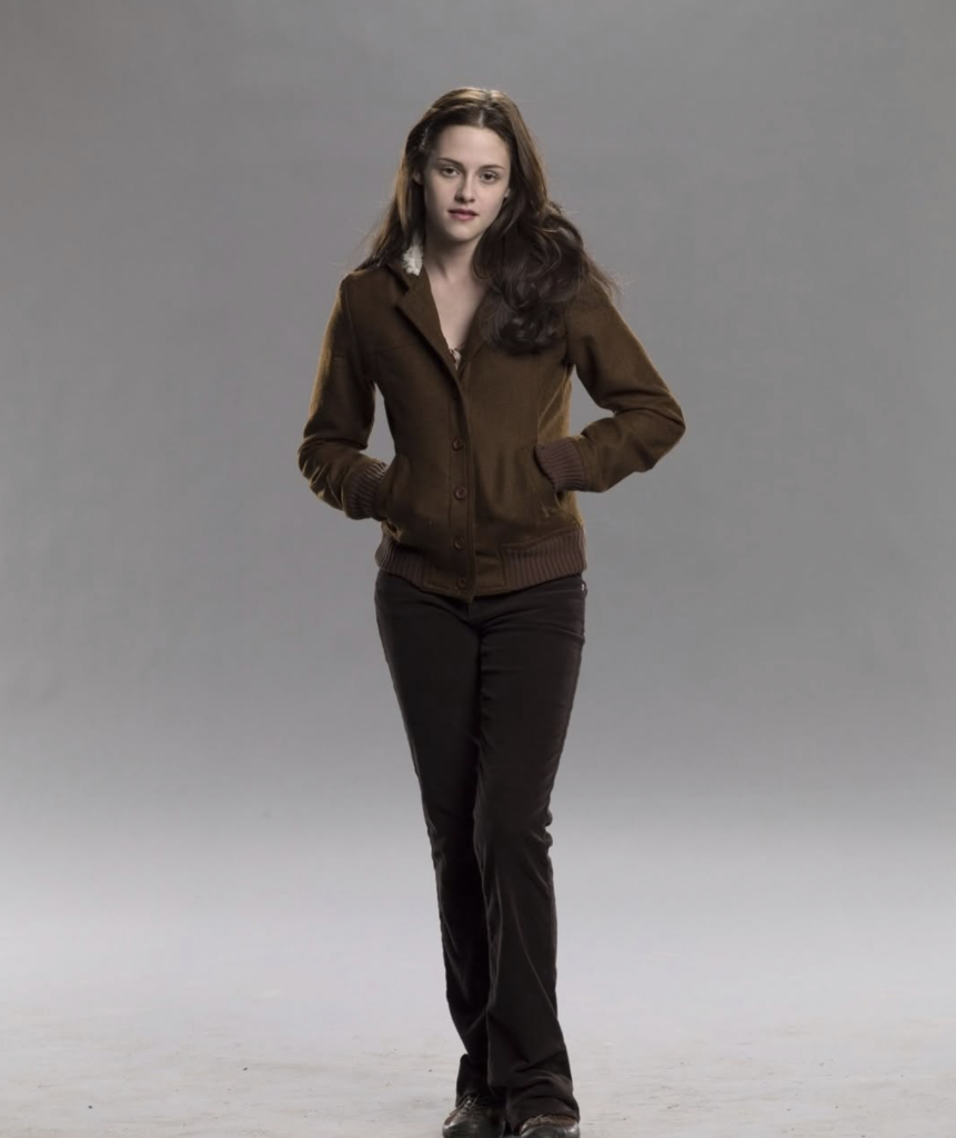 Kristen Stewart as Bella Swan, standing against a grey background wearing a brown jacket, dark pants, and a neutral expression.