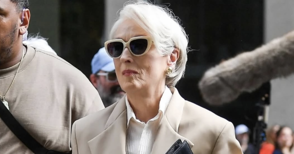 Hollywood actress Meryl Streep walking confidently in a beige oversized blazer, white shirt, and dark sunglasses, carrying a black leather clutch.