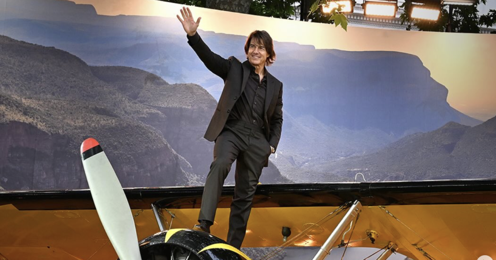 Actor Tom Cruise waving while standing on top of a yellow and black vintage biplane.