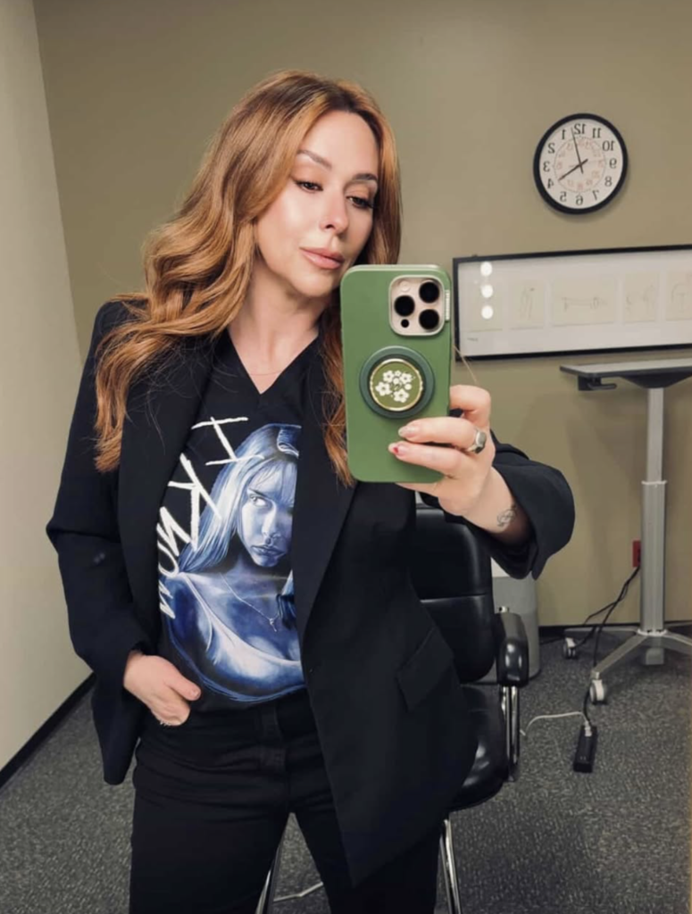 jennifer love hewitt with long wavy auburn hair taking a mirror selfie wearing a black blazer and graphic tee.