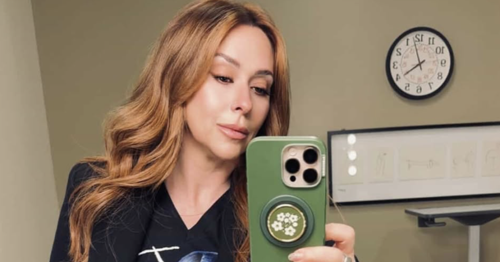 jennifer love hewitt with long wavy auburn hair taking a mirror selfie wearing a black blazer and graphic tee.