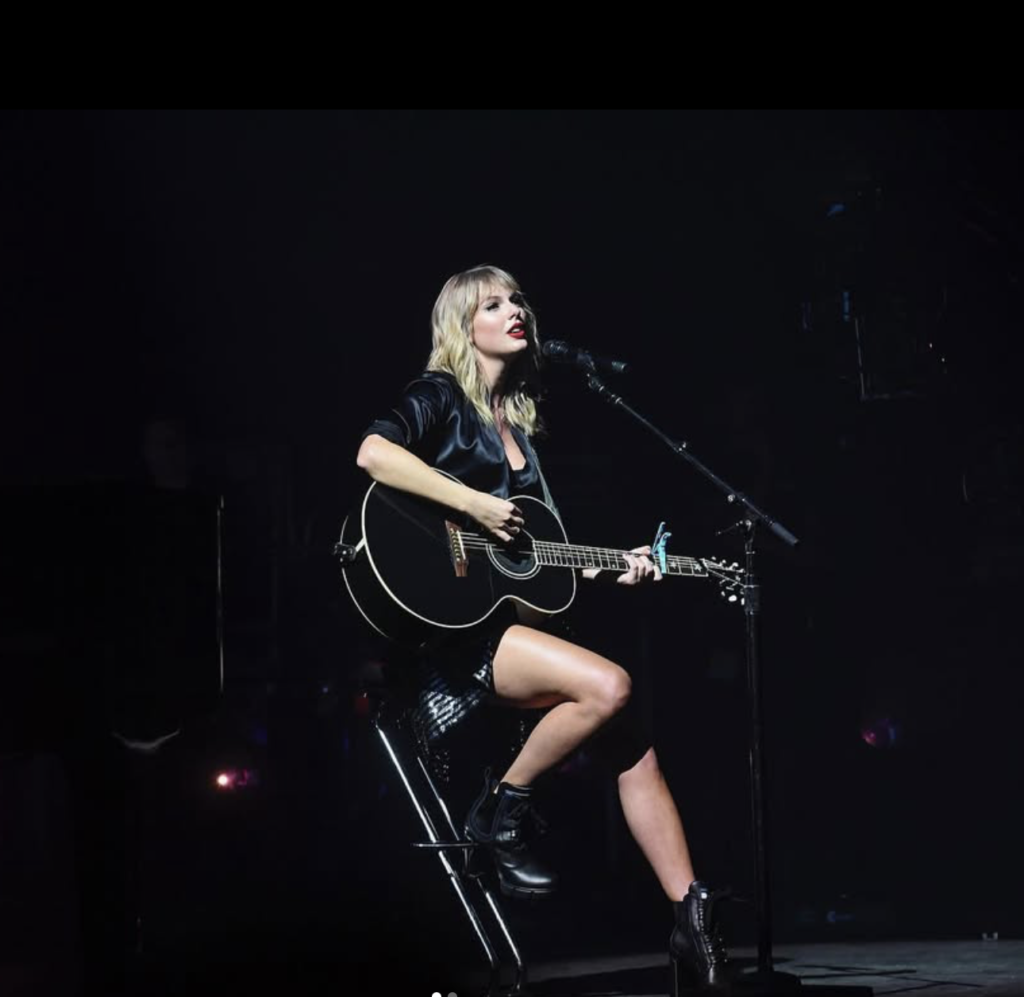 Taylor Swift performing on stage, sitting on a stool and playing a black acoustic guitar under a spotlight.