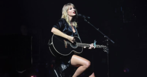 Taylor Swift performing on stage, sitting on a stool and playing a black acoustic guitar under a spotlight.