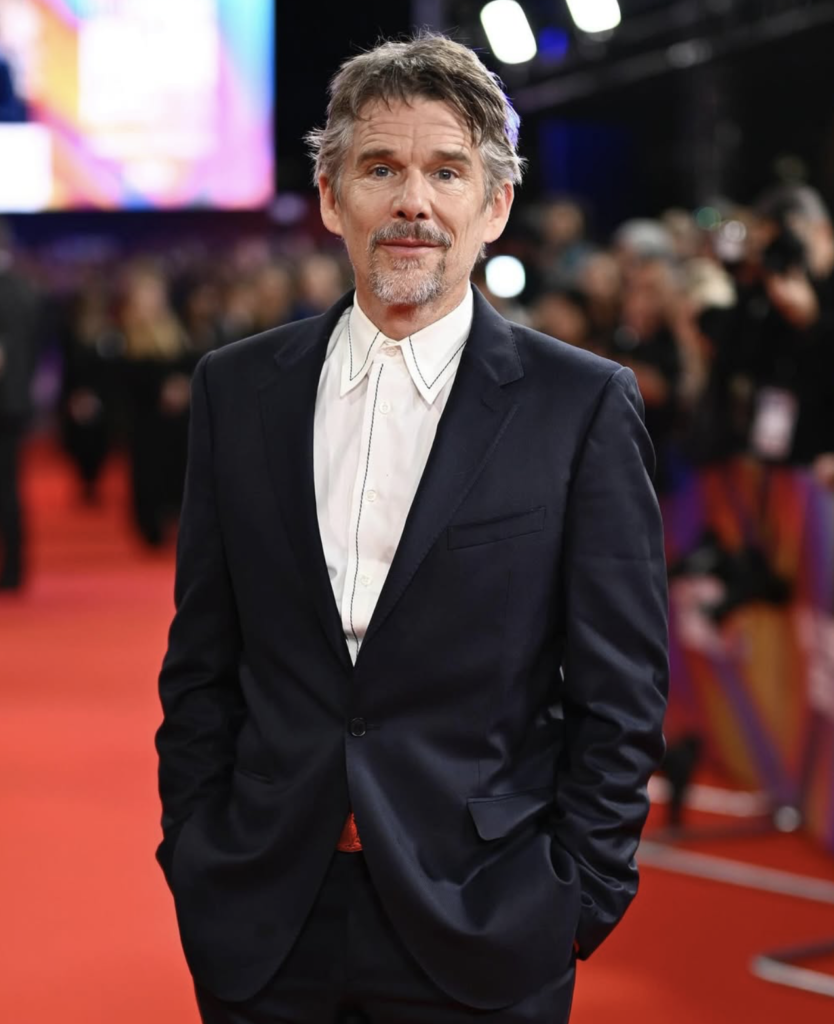  Ethan Hawke standing on a red carpet wearing a dark navy suit and a white shirt with black contrast stitching. He has messy gray hair and a goatee, looking directly at the camera with a subtle smile.