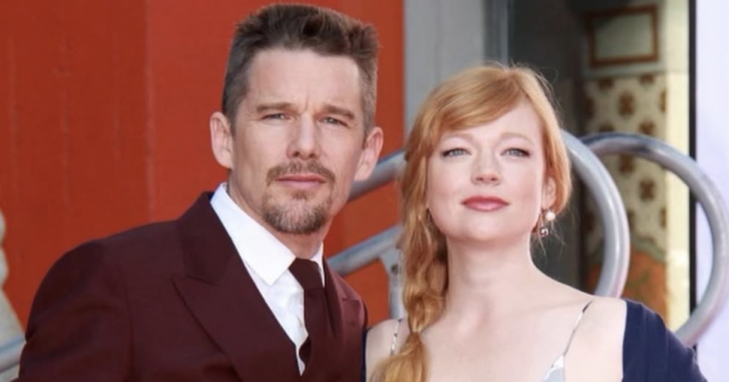 Ethan Hawke and Sarah Snook posing together at an event. Hawke wears a burgundy double-breasted suit with a tie, while Snook wears a silver floral dress with a navy cardigan and braided red hair.