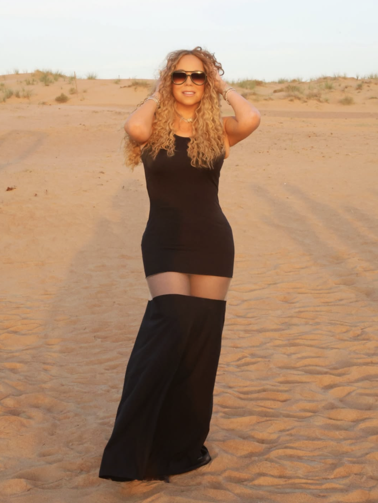 Mariah Carey posing in a desert at sunset, wearing a short black dress, oversized sunglasses, and unique, waist-high black boots.